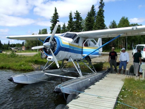 A seaplane with floats is docked by a wooden platform, with people nearby and trees in the background under a blue sky.