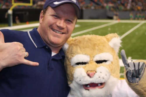 A smiling man poses with a mascot in a sports setting, both giving a thumbs-up gesture.