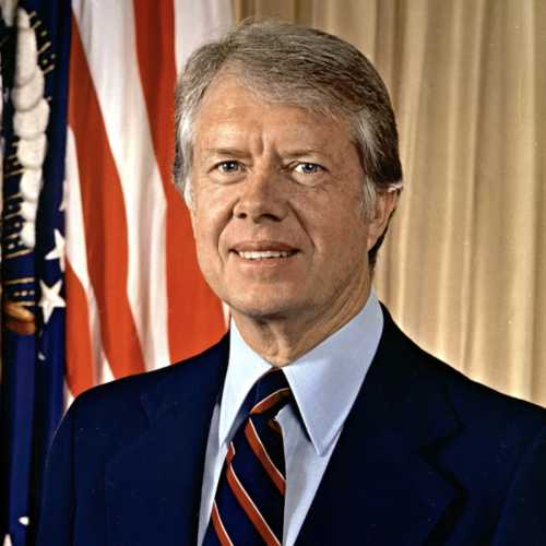 A man in a suit with a blue tie, smiling, stands in front of American flags and a beige backdrop.