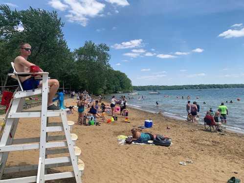 A lifeguard sits on a chair overlooking a busy beach with people swimming, sunbathing, and enjoying the sunny day.