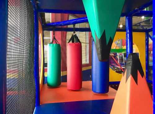 Colorful play area featuring hanging punching bags in red, green, and blue, with vibrant geometric shapes in the background.