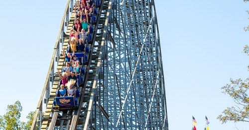 A wooden roller coaster with riders ascending a steep hill against a clear blue sky.