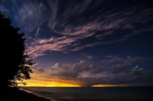 A serene night sky over a calm lake, with clouds illuminated by a distant glow and stars twinkling above.