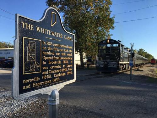 A historical marker for the Whitewater Canal with a train in the background, showcasing the canal's construction history.