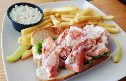 A lobster roll with lettuce, served with fries and a side of coleslaw on a white plate.