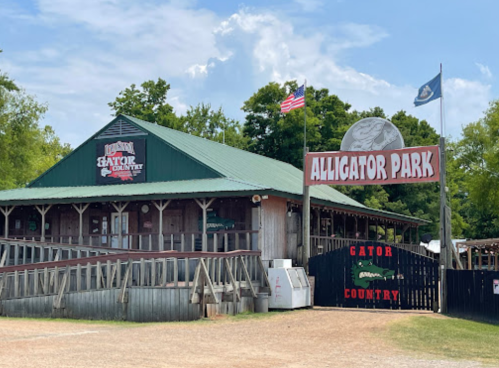 A rustic building with a green roof and signs for "Alligator Park" and "Gator Country," surrounded by trees and flags.