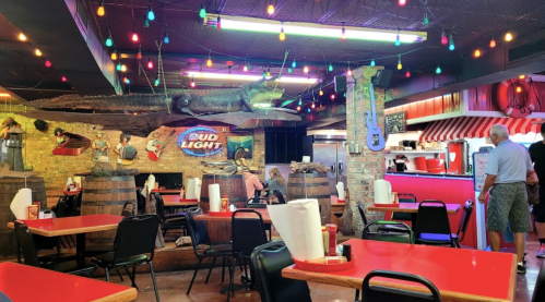 A lively restaurant interior with colorful lights, wooden barrels, and a large fish decoration on the wall.