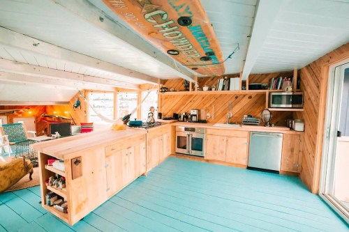 Bright, airy kitchen with wooden cabinets, blue floor, and surfboard decor, featuring modern appliances and cozy seating.