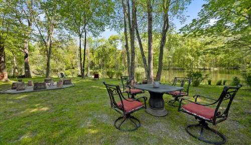 A serene outdoor setting with a round table and chairs by a pond, surrounded by lush greenery and trees.