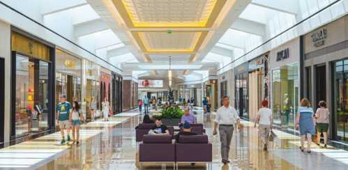 A bright, modern shopping mall with people walking, stores on either side, and seating areas in the center.