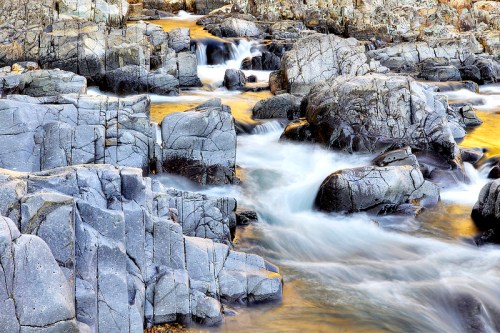 A serene river flows over smooth, gray rocks, creating gentle ripples and reflections in the water.