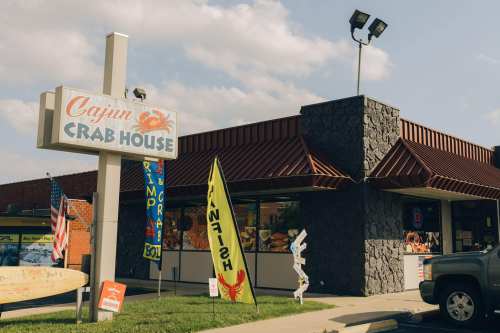 Exterior of Cajun Crab House with a sign, flags, and a stone facade on a sunny day.
