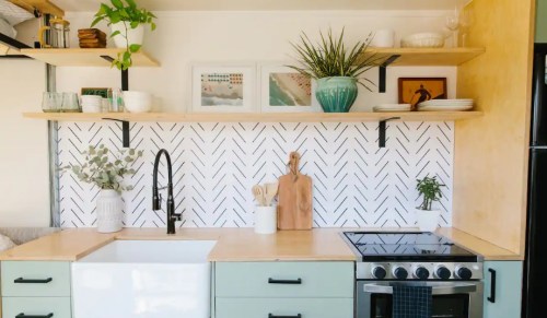 A modern kitchen with a farmhouse sink, stove, open shelves, and decorative plants against a herringbone-patterned backsplash.