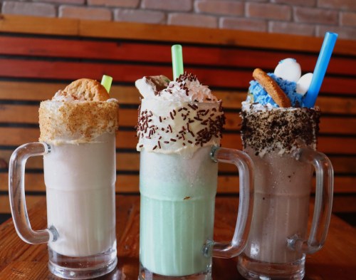 Three tall milkshakes with colorful toppings, including cookies, sprinkles, and whipped cream, on a wooden table.