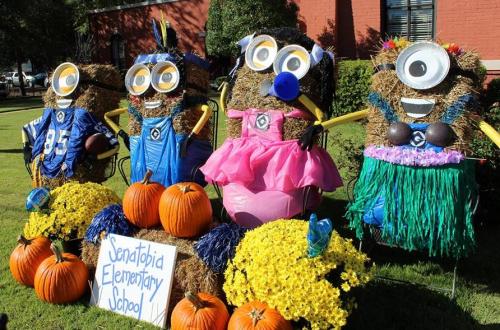 Four scarecrows dressed as Minions, with colorful outfits, surrounded by pumpkins and flowers outside Senatobia Elementary School.