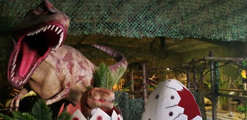A large dinosaur model with open jaws stands beside dinosaur eggs in a lush, green-themed environment.