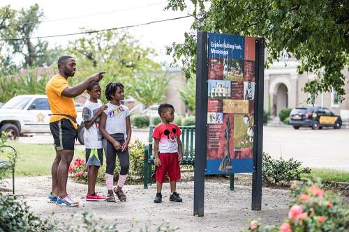 A man points at an informational sign while three children look on, surrounded by greenery and a park setting.