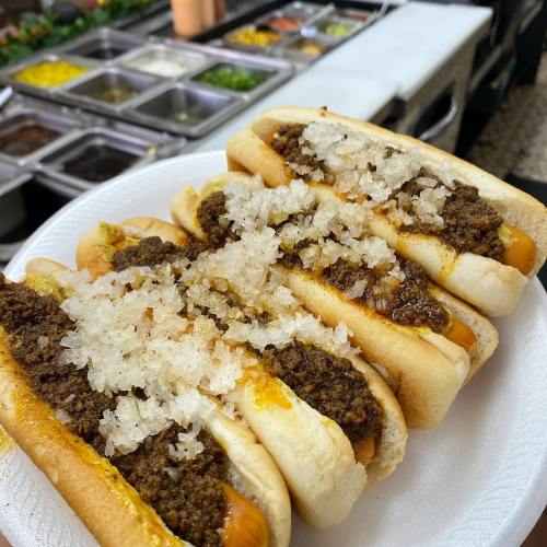 A plate of hot dogs topped with chili and chopped onions, set against a background of condiments and toppings.