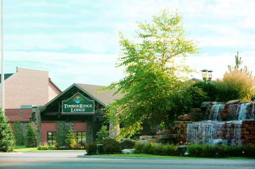 Timber Ridge Lodge with a waterfall and greenery in the foreground, set against a clear sky.