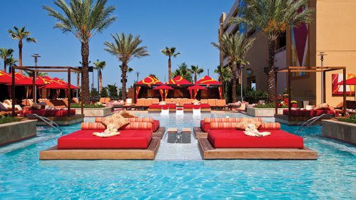 A luxurious pool area with red lounge beds, palm trees, and cabanas under a clear blue sky.