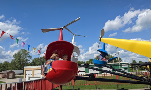 Children enjoy a colorful helicopter ride at an amusement park under a bright blue sky with fluffy clouds.