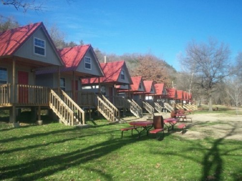 Row of cozy cabins with red roofs, surrounded by green grass and trees under a clear blue sky.