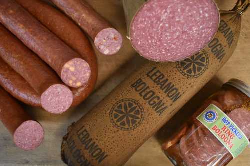 Various types of bologna, including sliced and whole, alongside a jar of pickled bologna on a wooden surface.