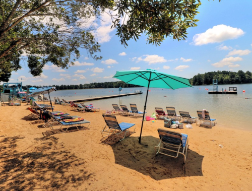 A sandy beach with lounge chairs and umbrellas, overlooking a calm lake under a bright blue sky with fluffy clouds.