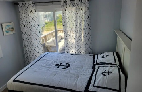 A cozy bedroom featuring a bed with anchor-patterned bedding and sheer curtains, overlooking a sunny outdoor area.