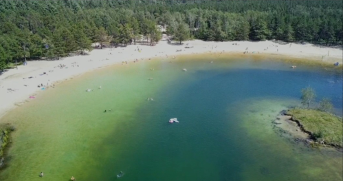 Aerial view of a beach with people swimming in a clear blue-green lake surrounded by trees.