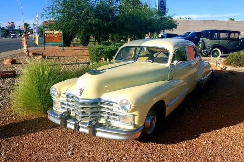 A vintage yellow car parked in a desert landscape with greenery and a clear blue sky in the background.