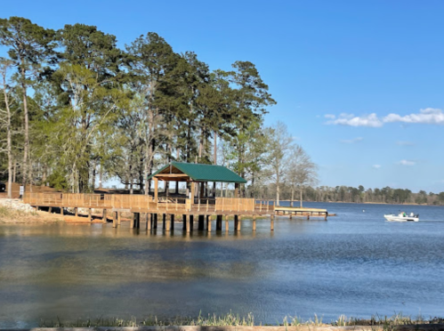 A wooden dock with a green-roofed gazebo extends over a calm lake, surrounded by trees under a clear blue sky.