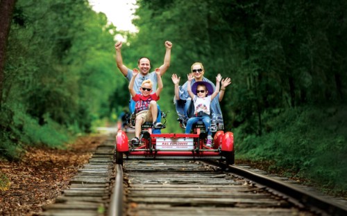 A family of four joyfully rides a pedal-powered cart on a scenic railway track surrounded by trees.