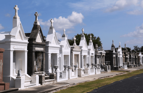 A row of ornate white mausoleums in a cemetery under a blue sky with scattered clouds.