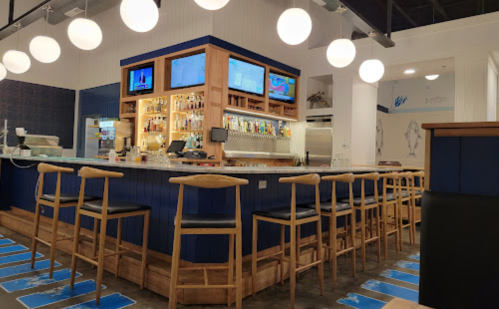 A modern bar with wooden stools, illuminated by round lights, featuring a variety of drinks and two screens.