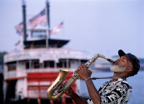 A musician plays saxophone in front of a riverboat, with American flags waving in the background.