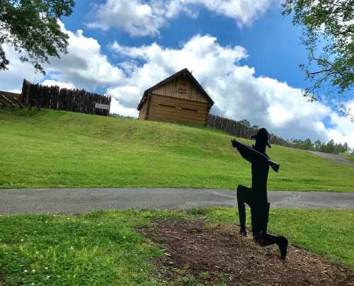 A silhouette of a kneeling figure with a sword in front of a grassy hill and a wooden structure under a blue sky.