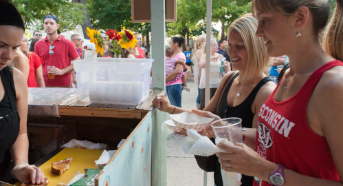 Two women smile while ordering food from a vendor at a busy outdoor event, with a crowd and greenery in the background.
