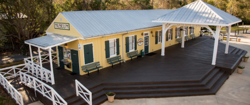 A yellow museum building with a metal roof, featuring a wooden deck and benches, surrounded by greenery.
