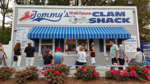 A busy clam shack with a blue-striped awning, people lined up at the window, surrounded by colorful flowers.