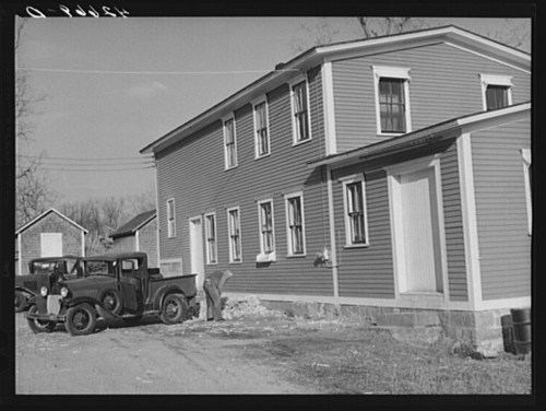 A vintage truck parked beside a two-story house with multiple windows, set in a rural area.