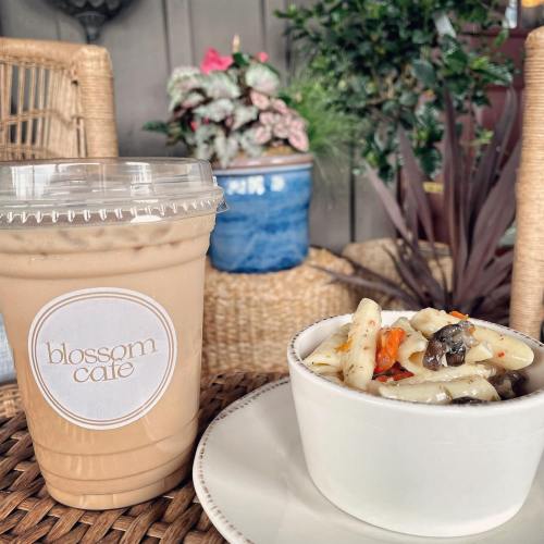 A cup of iced coffee next to a bowl of pasta salad, set on a woven table with plants in the background.