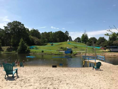 A sandy beach by a lake with green hills, blue slides, and people enjoying the water on a sunny day.