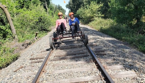 Two people in helmets ride a rail bike on a gravel track surrounded by greenery.