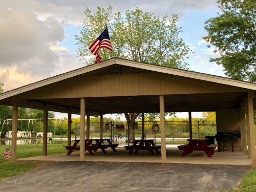 A covered picnic area with red tables, an American flag, and a grassy field in the background under a cloudy sky.