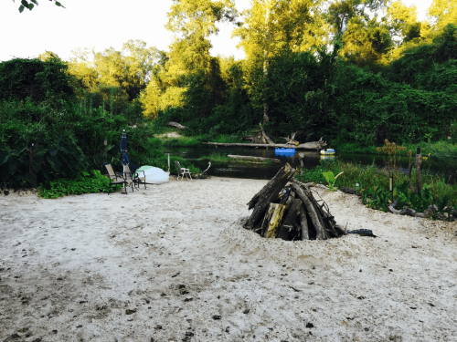 A sandy riverside scene with a fire pit, chairs, and lush greenery surrounding a calm waterway.
