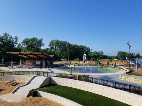 A sunny park featuring a splash pad, playground, and picnic area surrounded by green trees and blue sky.