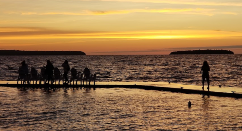 Silhouettes of people on a dock at sunset, with a calm sea and distant islands under a colorful sky.