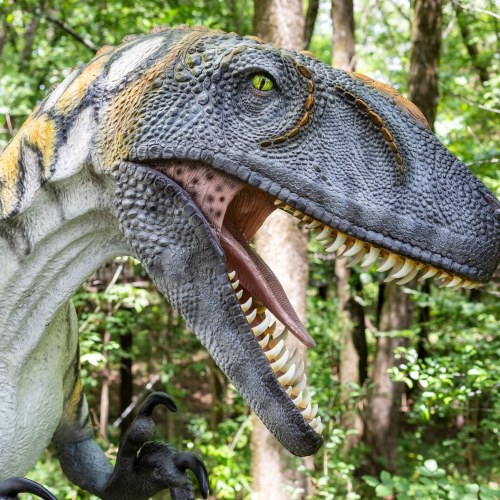 A close-up of a realistic dinosaur sculpture with textured skin and sharp teeth, set against a lush green forest background.