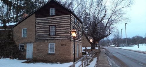 A historic two-story building with a mix of stone and wood, surrounded by snow and trees, along a quiet road.
